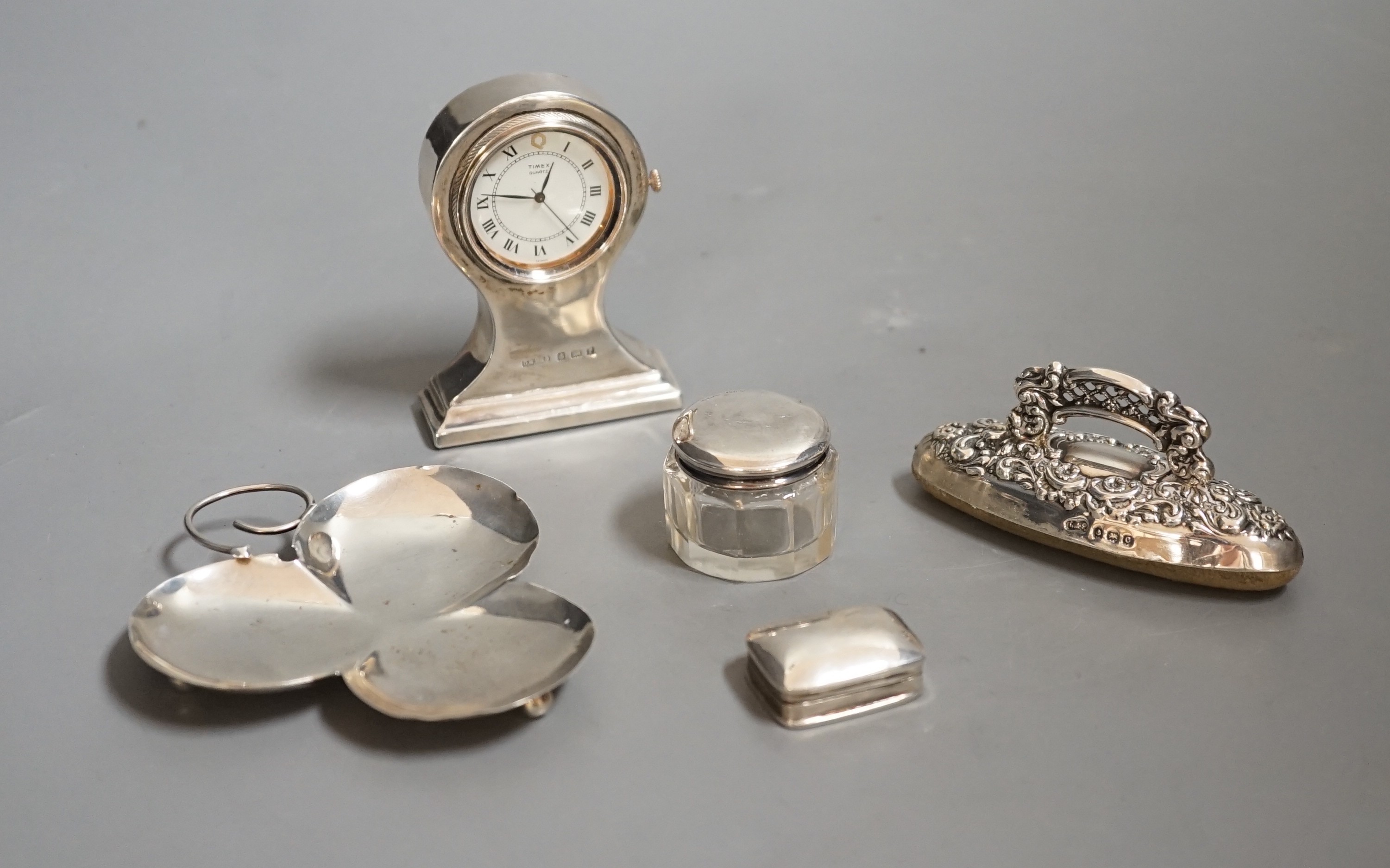An Edwardian miniature silver-cased mantel timepiece, snuff box, blotter, clover-shaped dish and silver-capped glass jar.